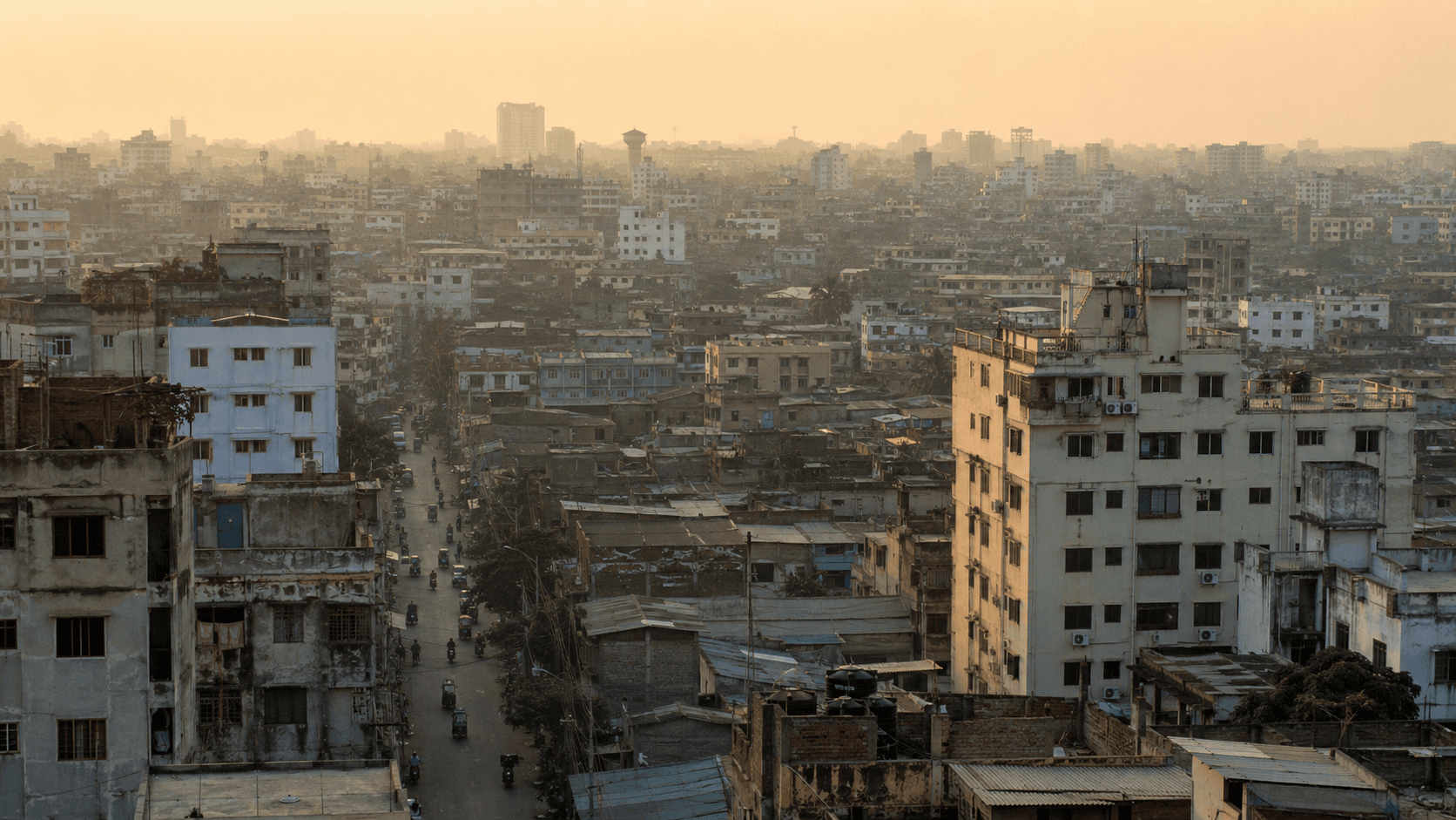 A dense Dhaka cityscape at golden hour, photographed from an elevated angle. Mid-rise concrete apartment buildings extend across the entire frame; lower-density tin-roof neighborhoods stretch to a hazy horizon where downtown towers rise. The sky is warm with late-afternoon sun but tinted yellow-brown from particulate pollution. Rooftop water tanks, power lines, and air-conditioning units visible throughout.