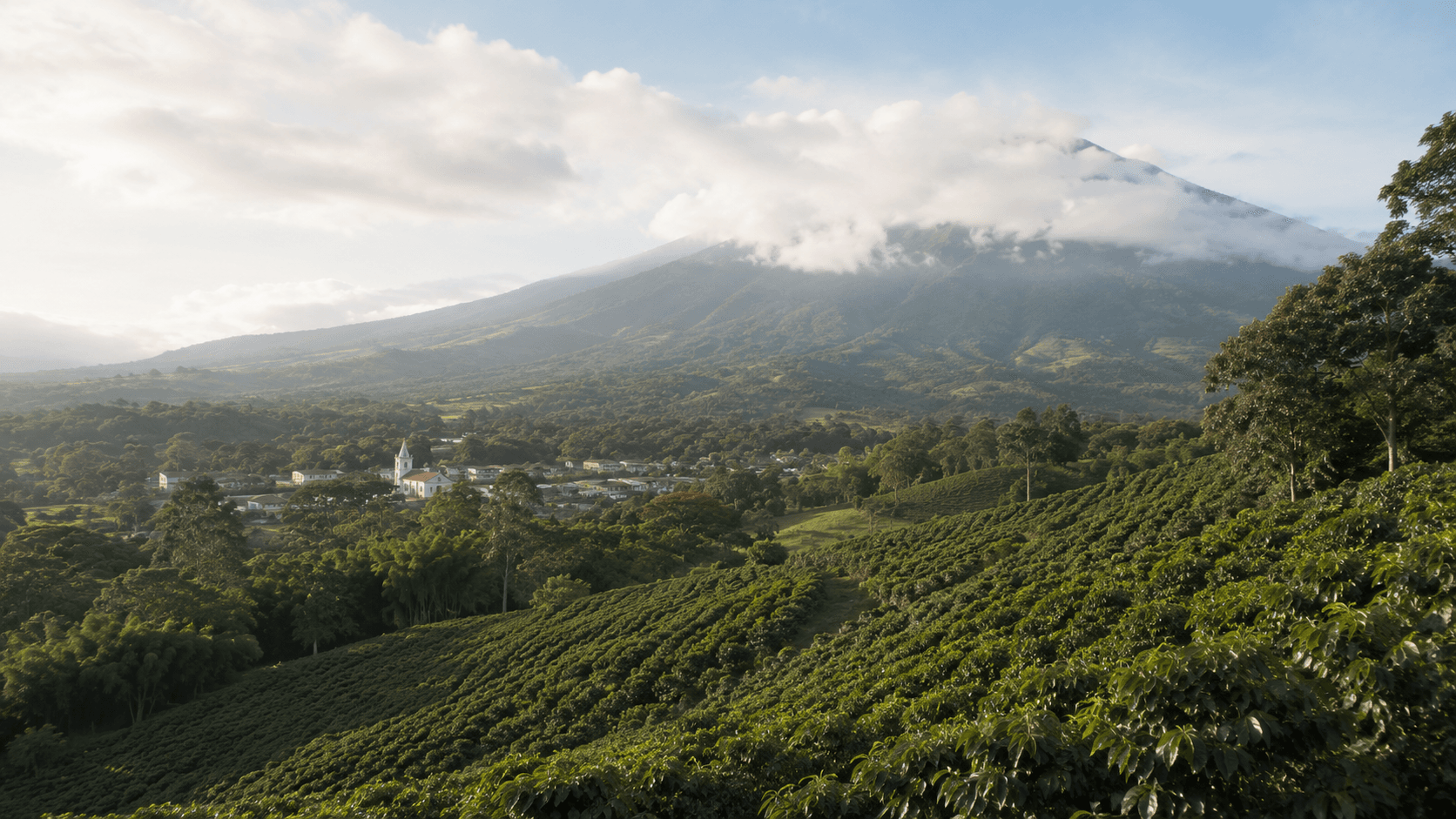 A green Costa Rican Central Valley landscape at morning: rolling coffee-covered hills, a small town with a white church among the trees, forested volcanic slopes rising into soft cloud behind. No people, no readable signage. The image evokes the pura-vida quality of life that draws retirees to Costa Rica's Pensionado visa, alongside the practical draw of access to the country's public-healthcare system.