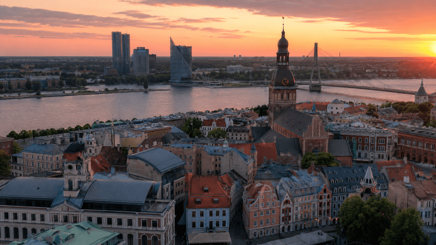 An aerial view of Riga, Latvia at sunset. Historic Old Town rooftops with red tiles dominate the foreground; the dark spire of Riga Cathedral rises in the center of the frame. The Daugava river crosses the middle distance, with modern glass towers of central Riga and a sweeping golden-orange sunset sky beyond.