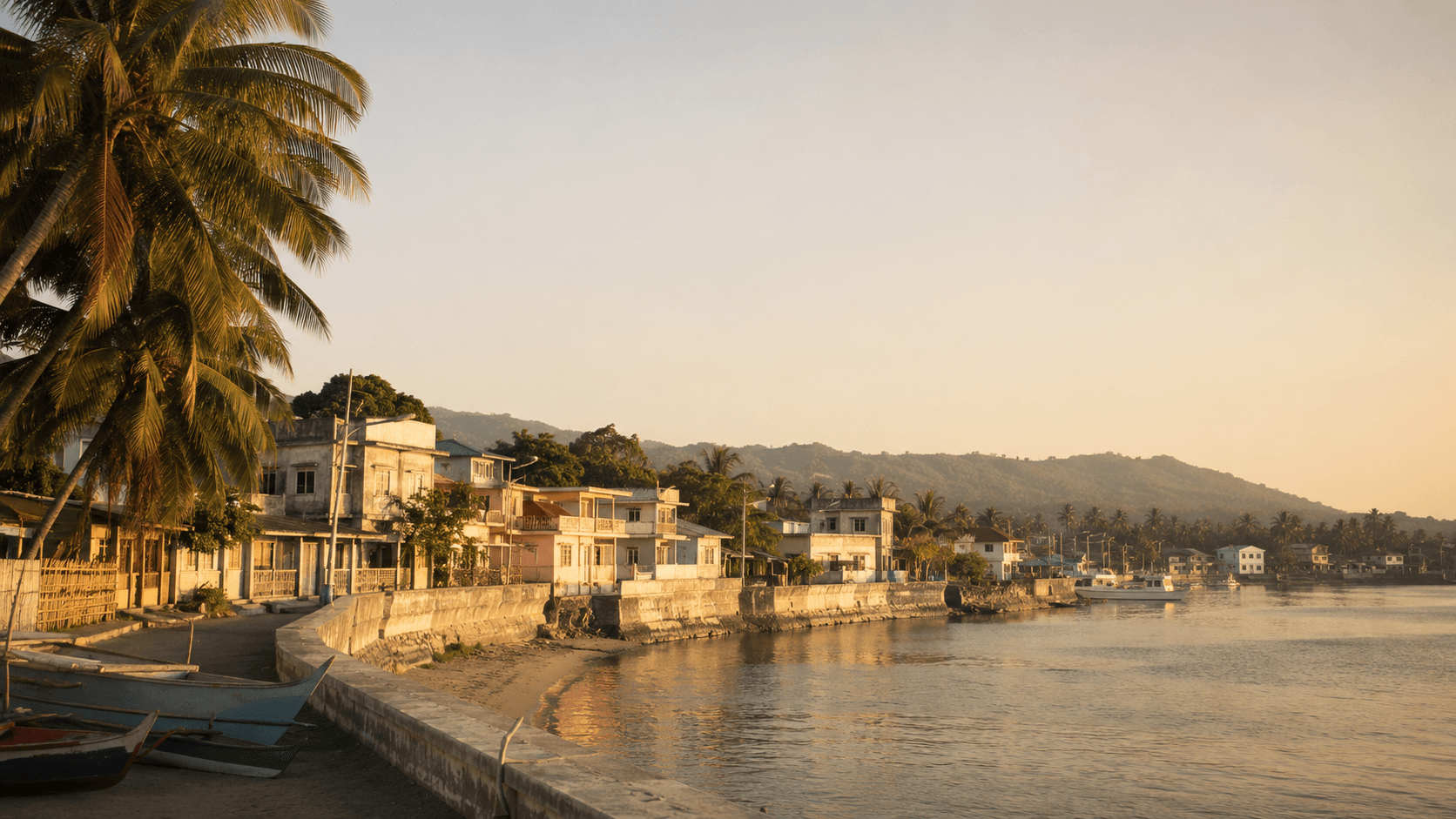 A quiet Philippine coastal town at golden hour: low pastel buildings along a calm waterfront, coconut palms leaning over a seawall, fishing bancas drawn up on the shore. No people, no readable signage. The image frames the Special Resident Retiree